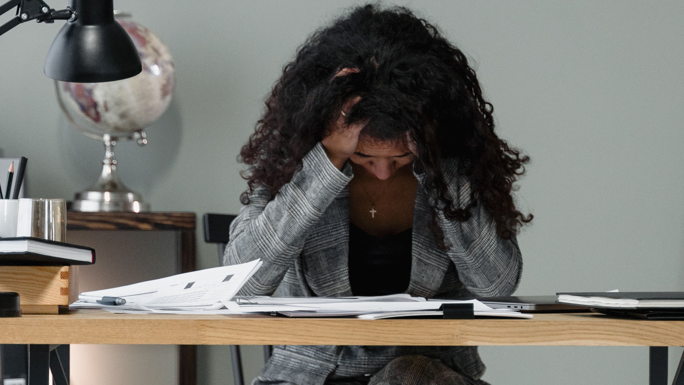 A woman with dark curly hair hold her head, looking down towards a dest piled with papers. She looks stressed.