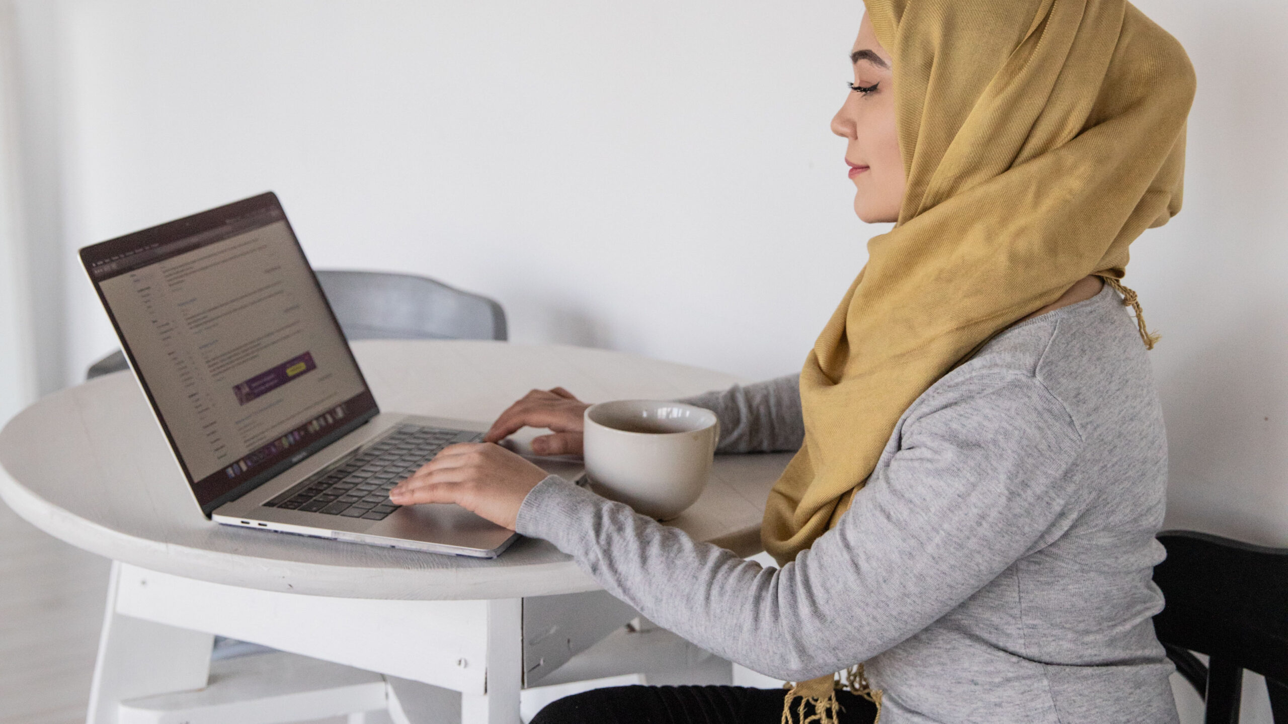 Woman sits at table using laptop. She wears a headscarf and has a coffee.