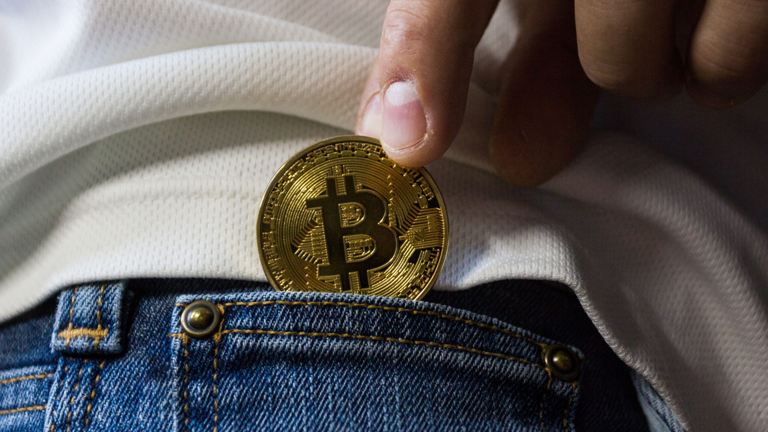 A hand places a coin with bitcoin symbol into jeans pocket.